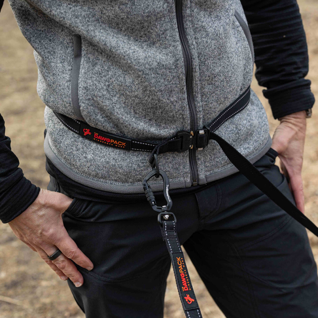 Person wearing a gray jacket with a black harness and leash, standing on a natural background.